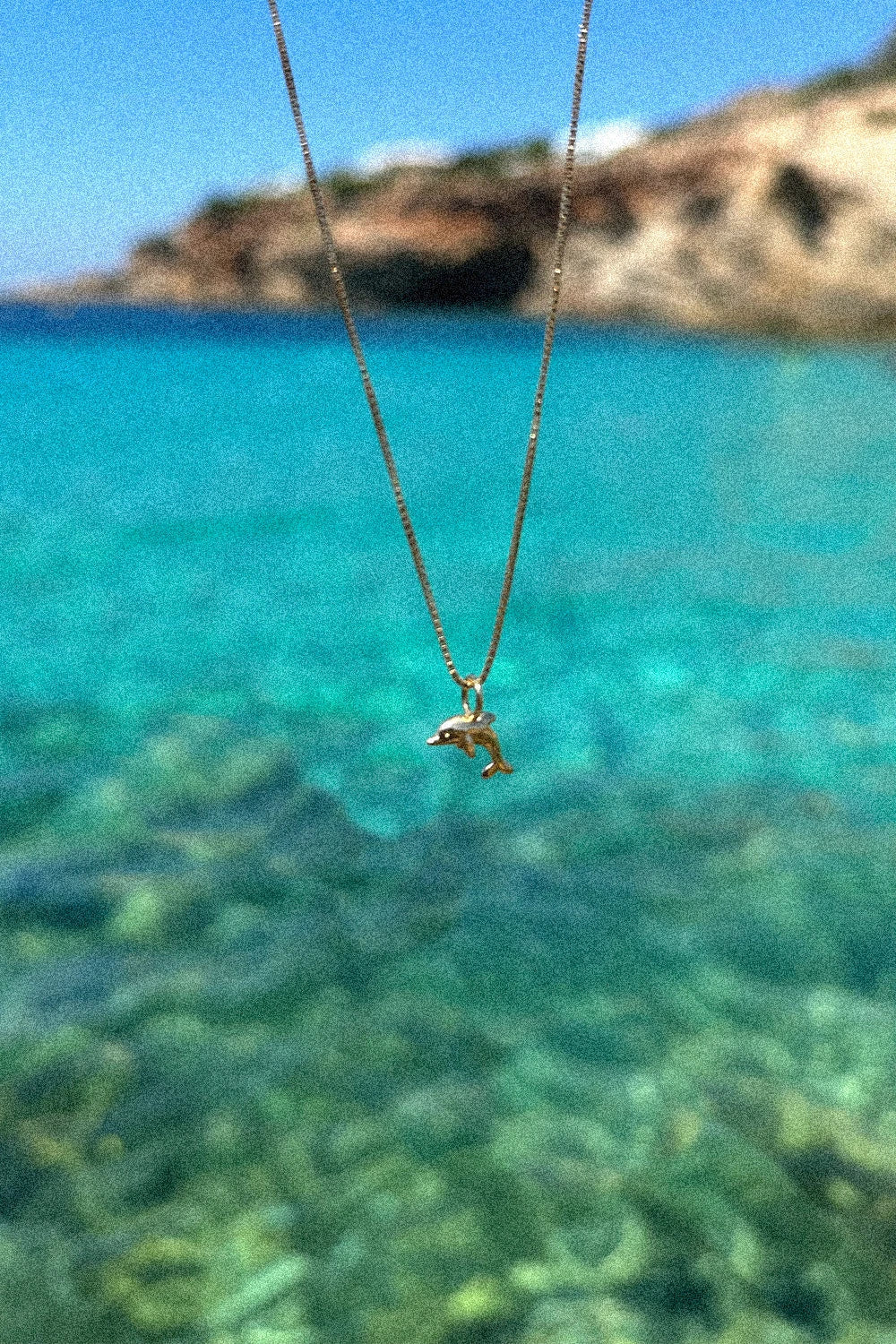 Dolphin Delfin-Anhänger aus 18 K recyceltem Echtgold von All About Articus, an einer Kette vor dem Meer