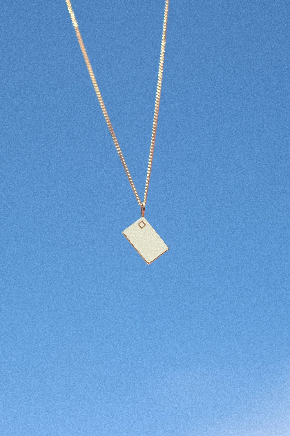 Postcard Paradiso Postkarten-Anhänger aus 18 K recyceltem Echtgold von All About Articus, an einer Kette vor blauem Himmel