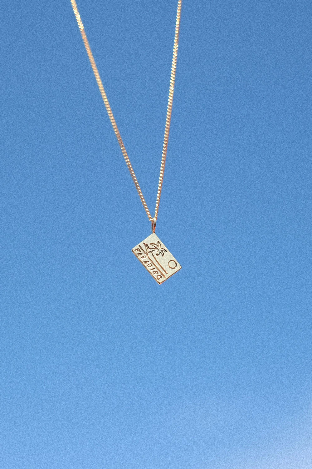 Postcard Paradiso Postkarten-Anhänger aus 18 K recyceltem Echtgold von All About Articus, an einer Kette vor blauem Himmel