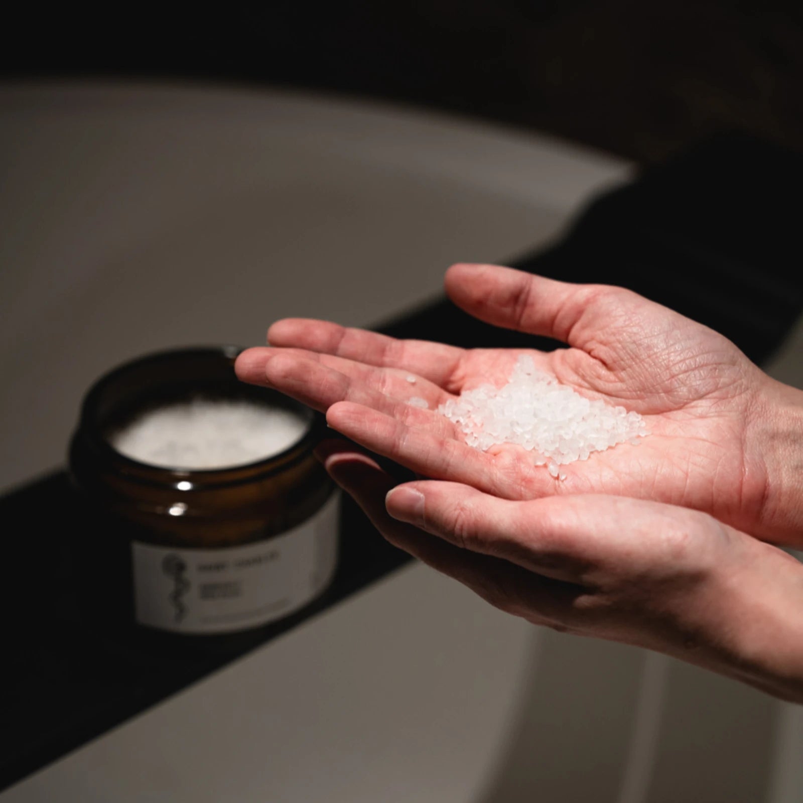 Hand mit SAINT CHARLES Badesalz Rising Heart vor geöffnetem Glastiegel am Badewannenrand