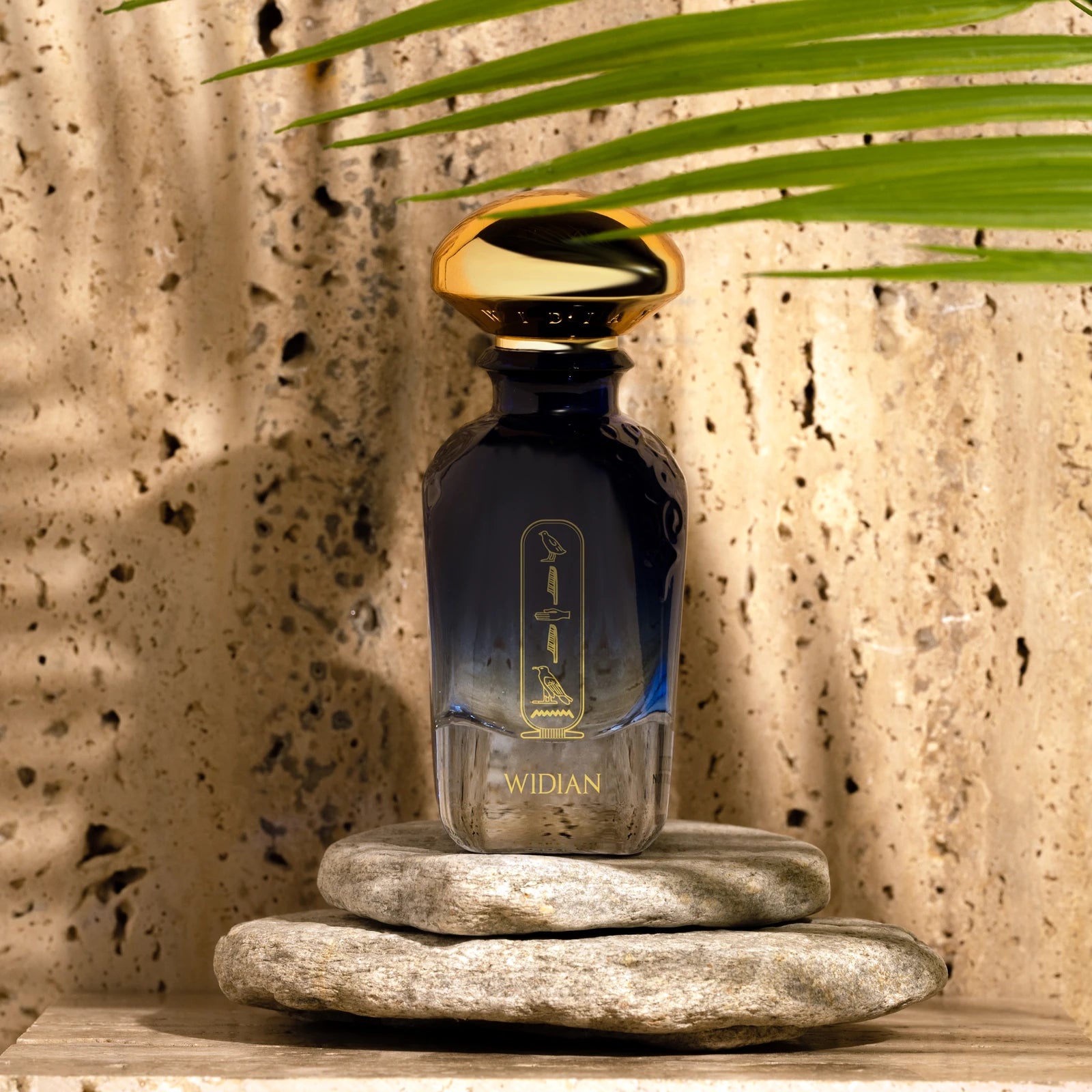 Widian Aswan bottle on stones in front of a sunlit travertine wall with a palm leaf